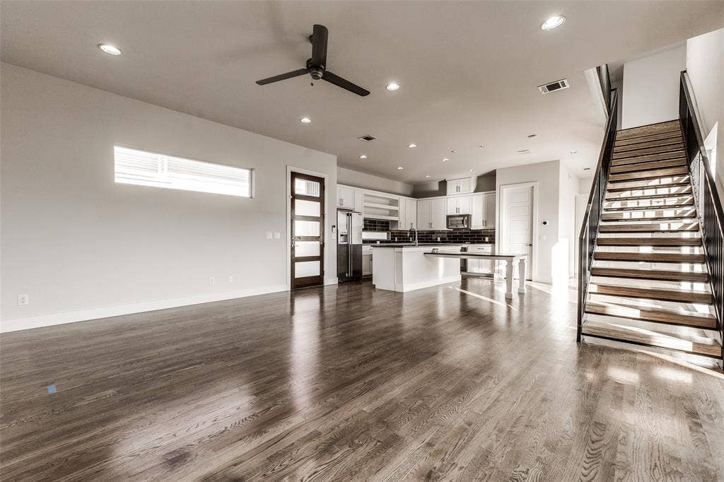 Open-concept interior featuring wood-finish flooring, recessed lighting, a modern staircase with metal handrails, a kitchen with white cabinetry, and a multi-panel glass door