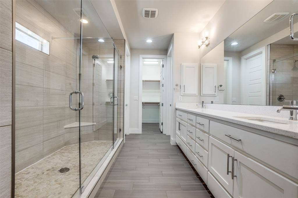 Bathroom featuring a glass-enclosed shower with tiled walls and a stone-style shower floor, double vanity with white cabinetry, quartz countertop, and large-format tile flooring