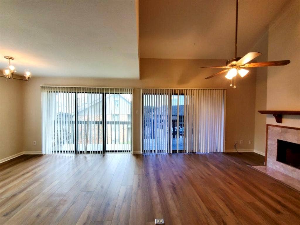 Vaulted ceiling featuring a central lighted ceiling fan, wood-finish flooring, dual sliding glass doors with vertical blinds, and a tile-surround fireplace with a wood mantel