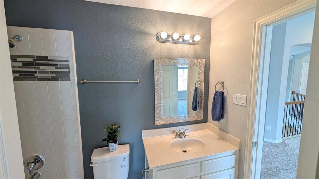 4221 Crestfield Drive, Richardson, TX 75082 Bathroom featuring a white vanity with a composite countertop, an integrated sink, a framed wall mirror, four-bulb vanity lighting, and a shower with tile surround and decorative horizontal accent inlay