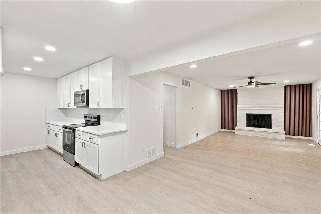 Kitchen featuring white shaker-style cabinetry, solid-surface countertops, and stainless steel appliances, accented by wood-finish flooring and a white painted brick fireplace