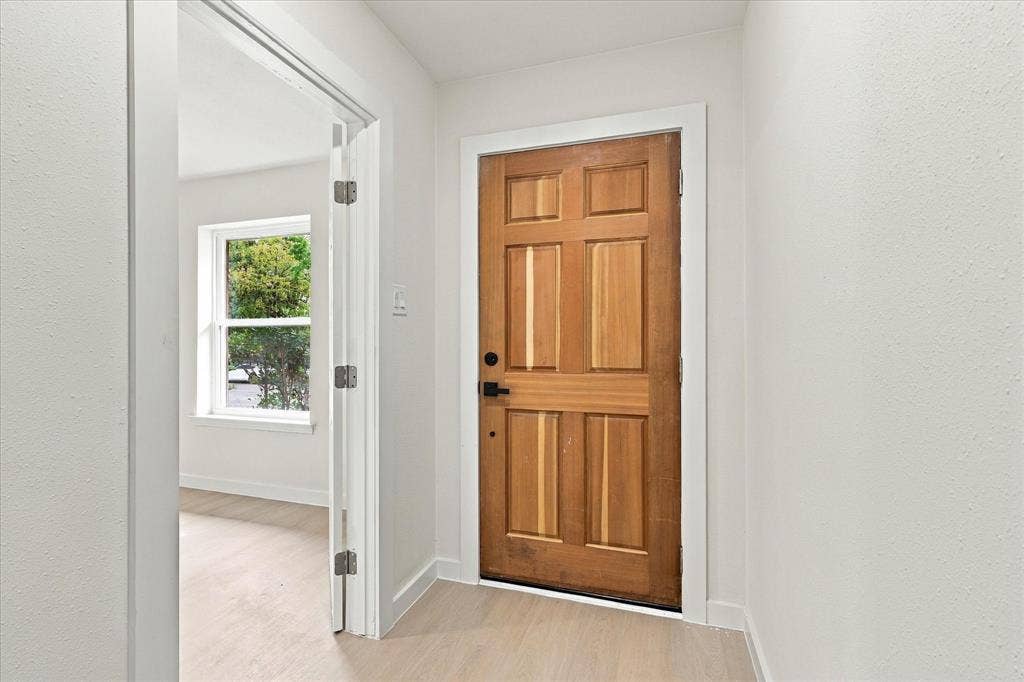 Six-panel natural wood entry door featuring matte black hardware, light-toned wood-finish flooring, and crisp white wall finishes