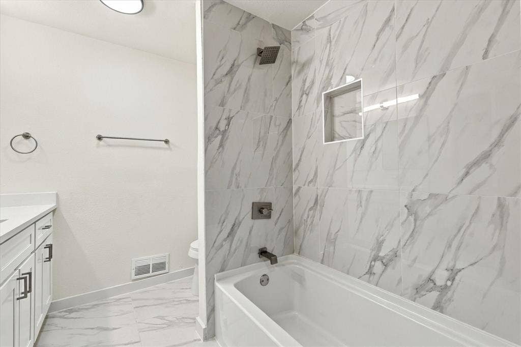 Bathroom featuring large-format veined tile surround, recessed wall niche, soaking tub, vanity with solid-surface countertop, and matte black fixtures