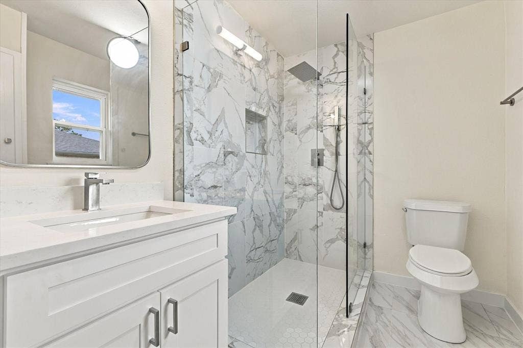 Bathroom featuring a stone-patterned tile walk-in shower with a built-in recessed niche, rainfall showerhead, glass enclosure, and white vanity with a stone countertop
