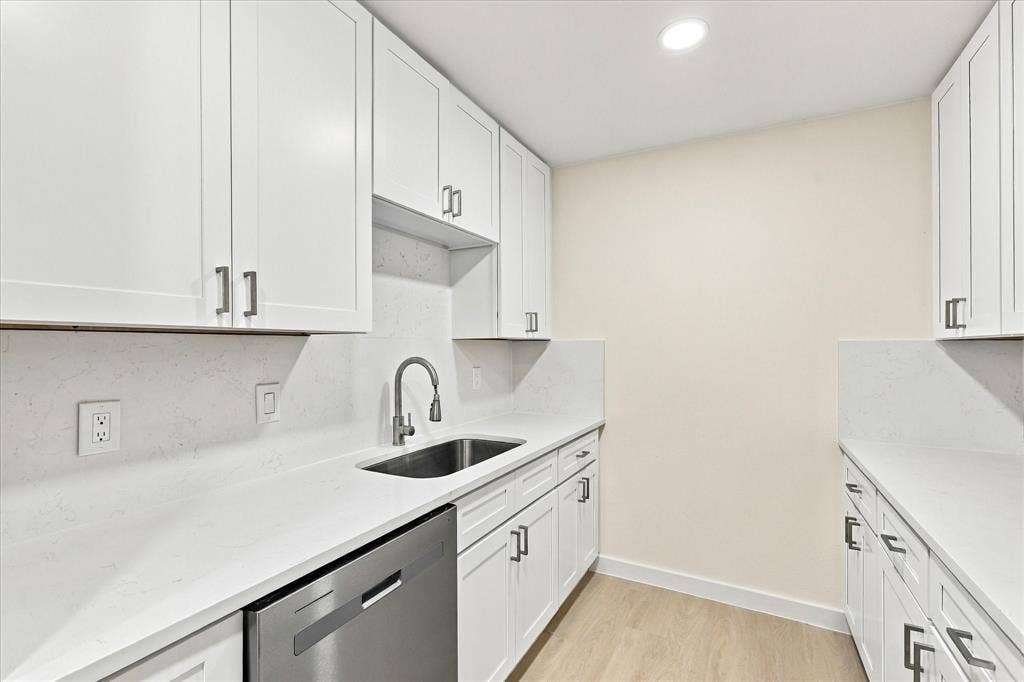 Shaker-style white cabinetry, solid-surface countertops with integrated backsplash, stainless steel under-mount sink with high-arc faucet, stainless steel dishwasher, and light-toned wood-finish flooring