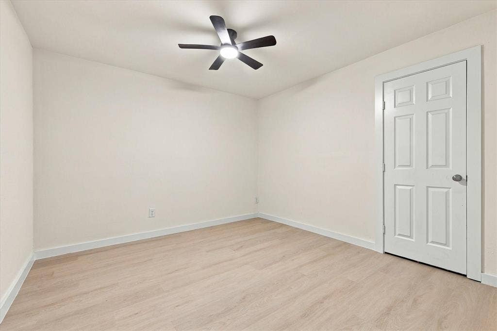 Neutral-toned interior space featuring light wood-finish flooring, a six-panel interior door, and a five-blade ceiling fan with integrated lighting
