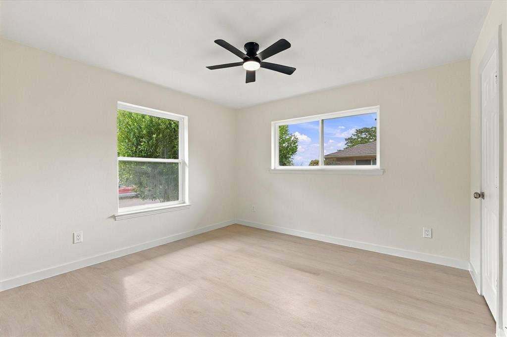 Neutral-toned interior space featuring light wood-finish flooring, two large windows, and a matte black ceiling fan with an integrated light fixture