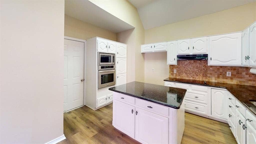 Kitchen featuring a vaulted ceiling, wood-finish flooring, white cabinetry, dark stone countertops, and a built-in double oven