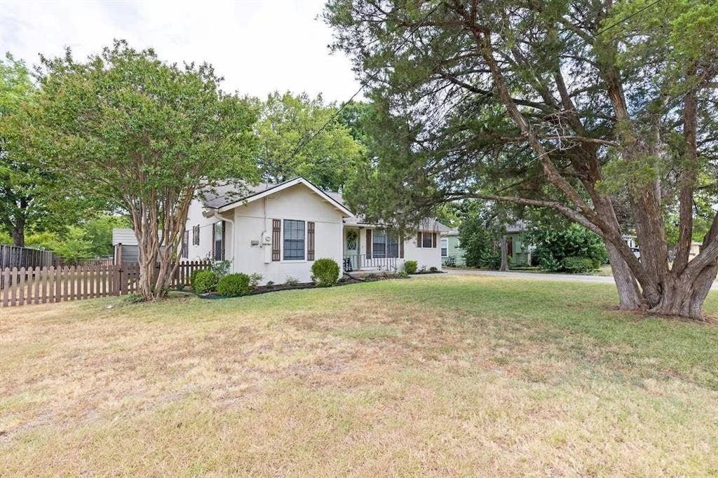 1101 N Waddill Street, McKinney, TX 75069 Single-story residence featuring light-toned exterior siding, louvered shutters, mature trees, and a wooden perimeter fence