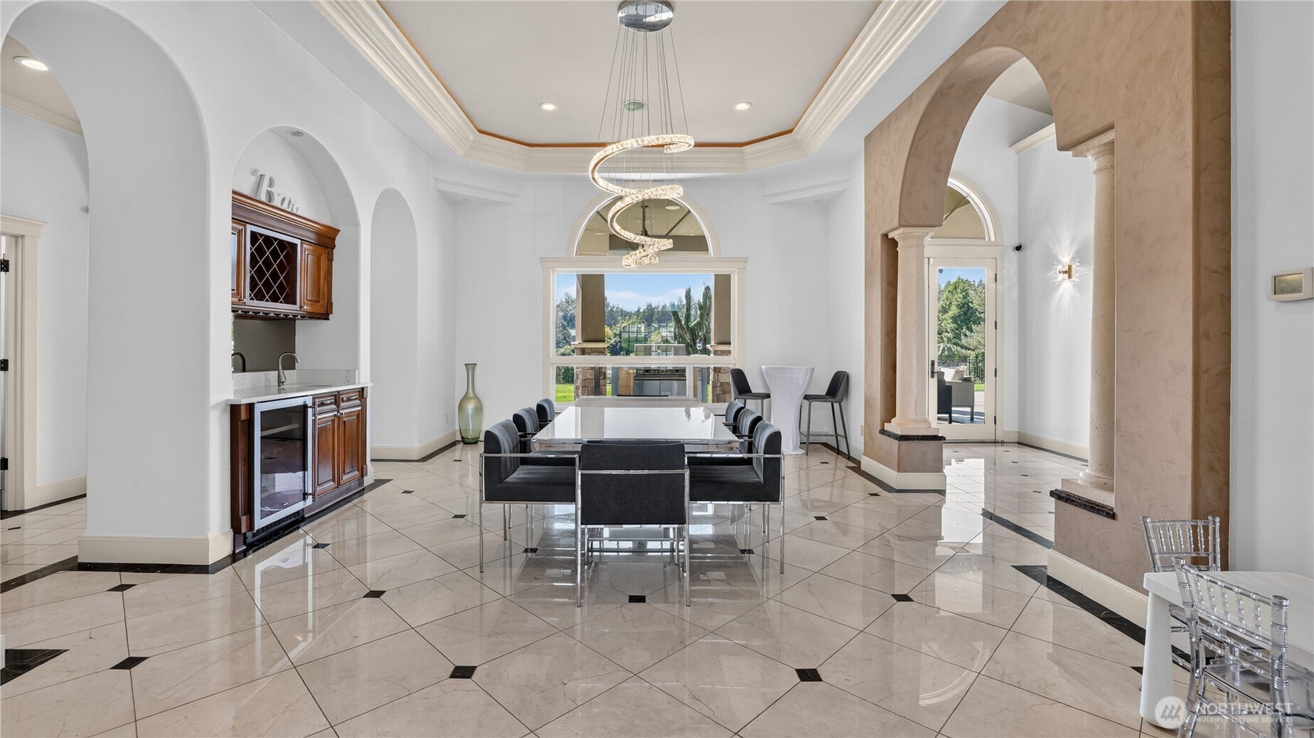 This exquisite formal dining room blends architectural grandeur with modern sophistication. Framed by graceful archways and stately columns, the space features polished marble floors and a coffered ceiling with elegant crown molding. A sculptural spiral chandelier serves as a stunning centerpiece above the sleek glass dining table, creating a perfect balance of artistry and light. The built-in bar with rich wood cabinetry and wine storage adds convenience and charm, ideal for entertaining. Expansive windows capture serene golf course views and fill the room with natural light, while the open flow to the patio invites seamless indoor-outdoor gatherings. Every detail reflects refined taste and timeless design.