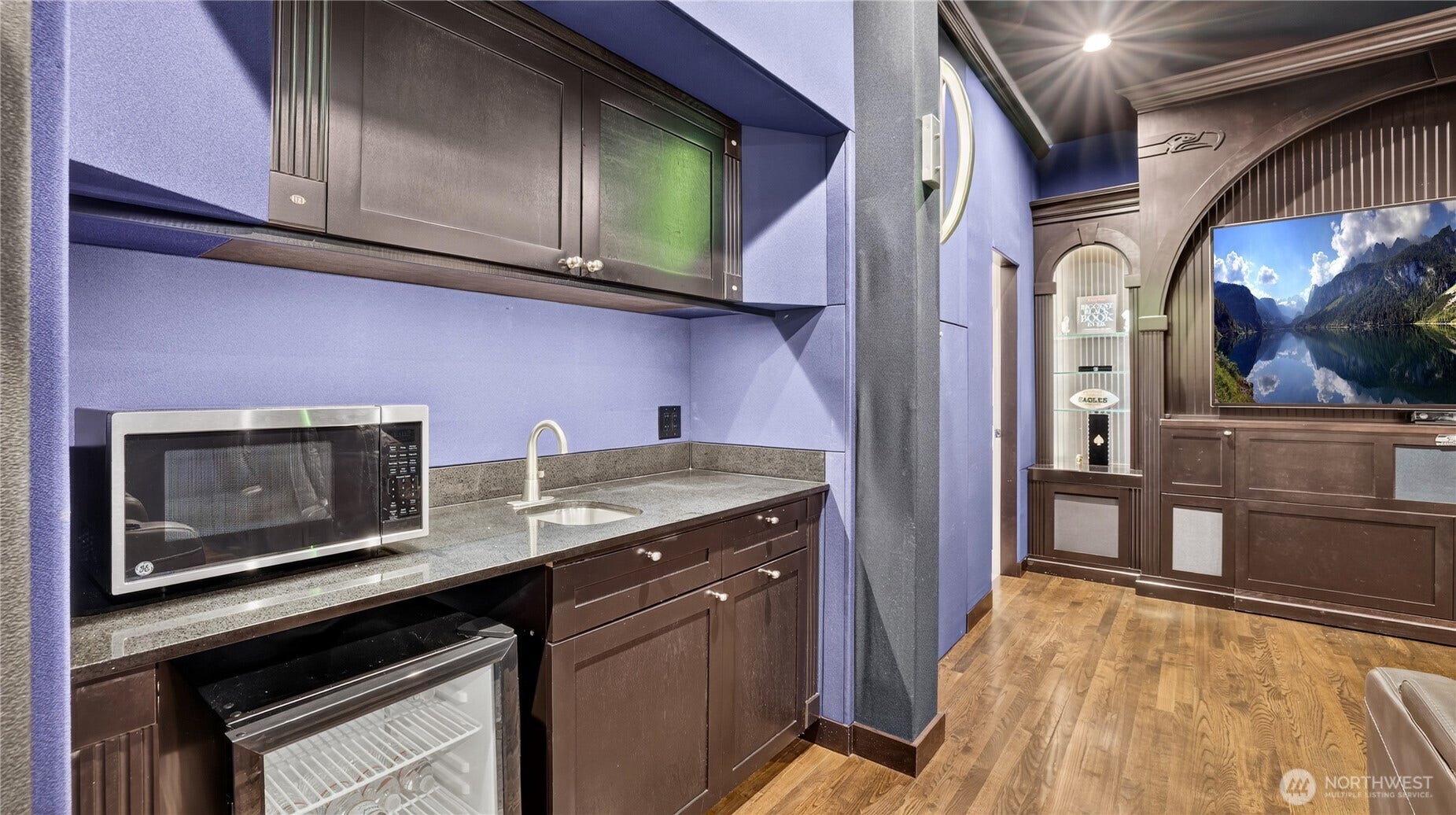 This stylish wet bar enhances the theater experience with sleek dark cabinetry, granite counters, and stainless-steel appliances  including a built-in microwave, mini-fridge, and sink. Perfect for serving refreshments during movie nights or game days, it adds both convenience and sophistication to the space.