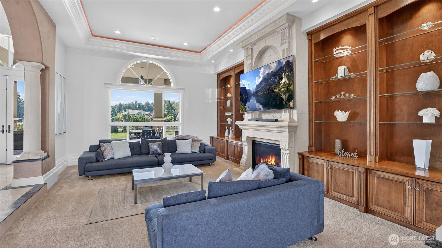 ophisticated and serene, this formal living room exudes timeless elegance with its soaring coffered ceilings, crown molding, and abundant natural light. Centered around a stately stone fireplace, the space is framed by custom wood built-ins that showcase refined craftsmanship and warm tones. Plush seating in soft blue hues invites conversation, while the large arched window perfectly captures views of the manicured grounds and outdoor entertaining area. Every detailfrom the recessed lighting to the symmetrical layoutcreates a harmonious balance of luxury and comfort, making this an ideal setting for both intimate gatherings and grand entertaining.