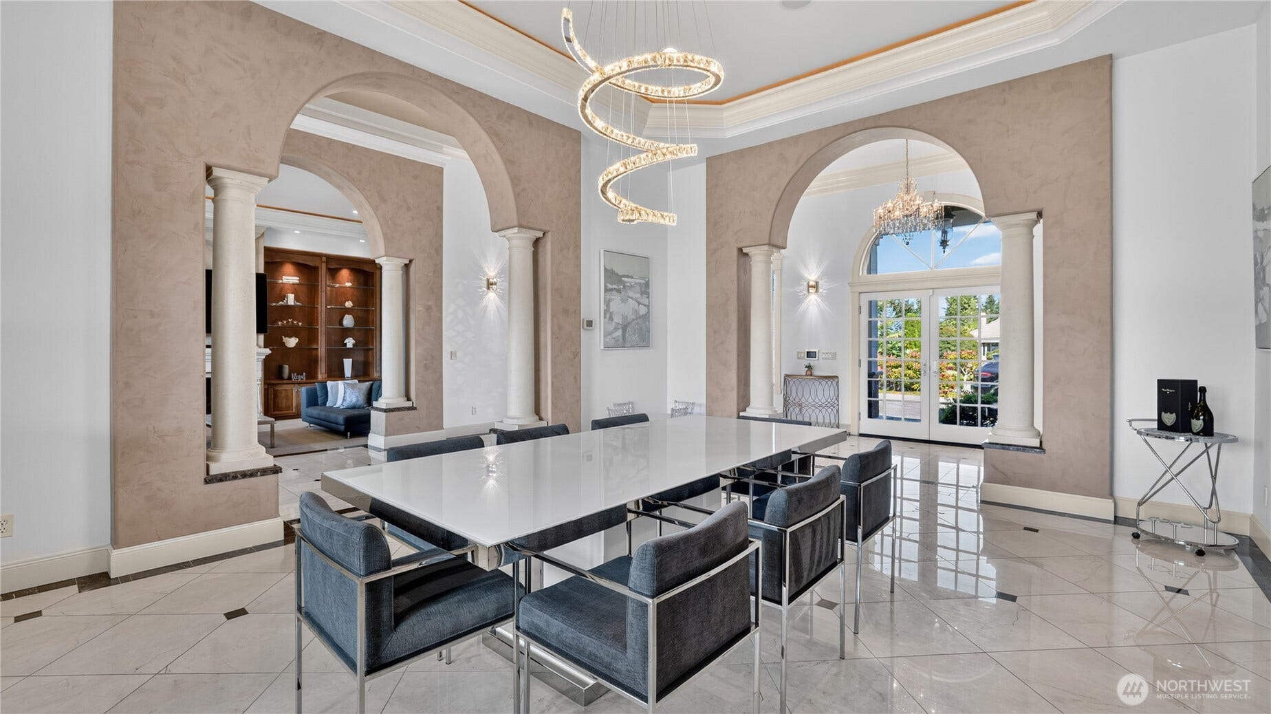 Formal Luxury Dining Room    This breathtaking formal dining room blends architectural grandeur with modern sophistication. Framed by arched columns and soaring ceilings, the space is anchored by a striking sculptural chandelier that adds a contemporary flourish. Polished marble floors and elegant wall finishes create a refined backdrop for gatherings, while natural light pours through the arched French doors, highlighting the seamless flow to adjoining living spaces. A perfect setting for elegant entertaining and memorable occasions.