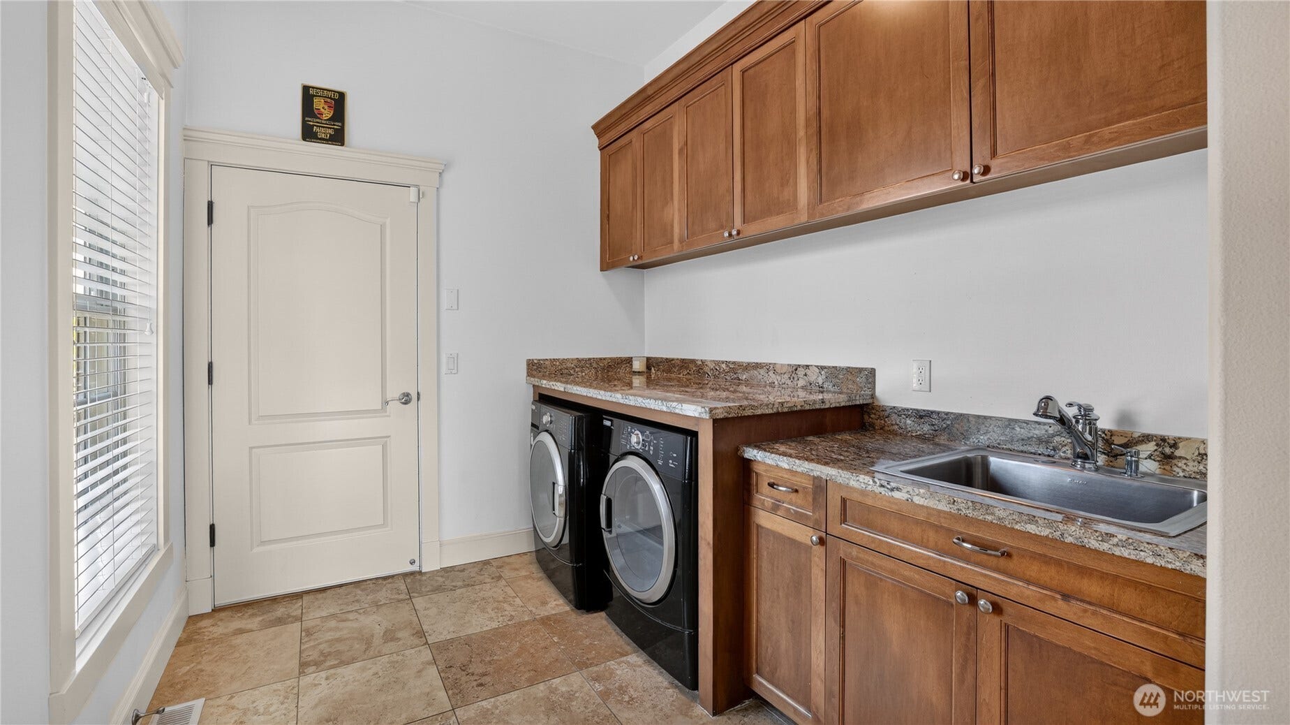 One of two beautifully appointed laundry rooms, this space combines practicality with refined design. Rich wood cabinetry offers generous storage above and below, complemented by polished granite countertops that provide ample workspace. A deep utility sink and modern front-loading washer and dryer make everyday tasks effortless. Natural light filters through the window, highlighting the warm tones of the tile flooring and the clean, crisp aesthetic. Thoughtfully designed for convenience and style, this laundry room reflects the same level of craftsmanship found throughout the home.