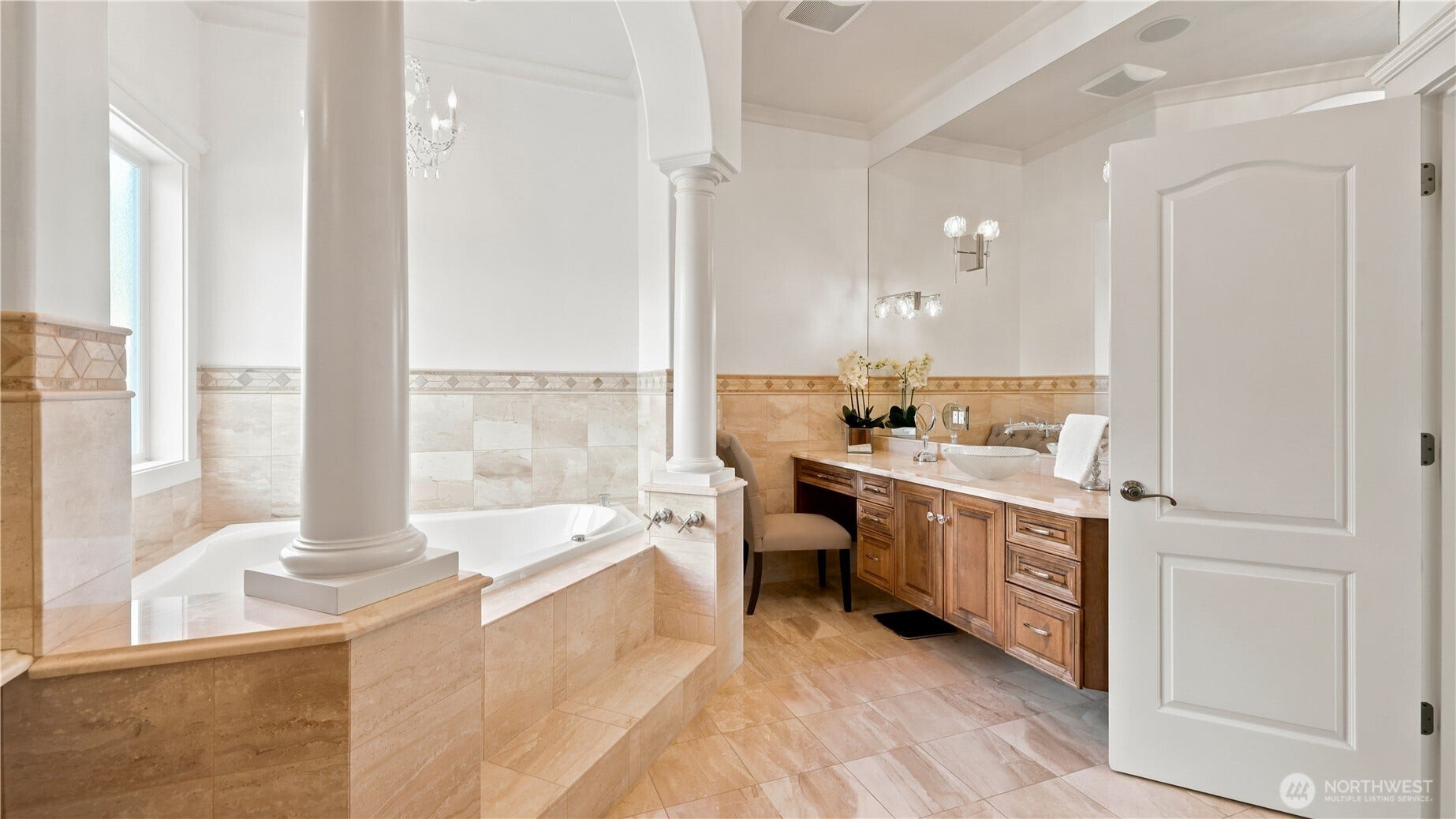 Primary Bathroom in Suite    This elegant primary bathroom exudes timeless sophistication with its arched columns, marble tile, and chandelier lighting. A deep soaking tub anchors the space, complemented by a custom vanity with rich wood cabinetry and a vessel sink. Thoughtfully designed for both beauty and function, the room offers a serene, retreat-style atmosphere that perfectly balances luxury and comfort.