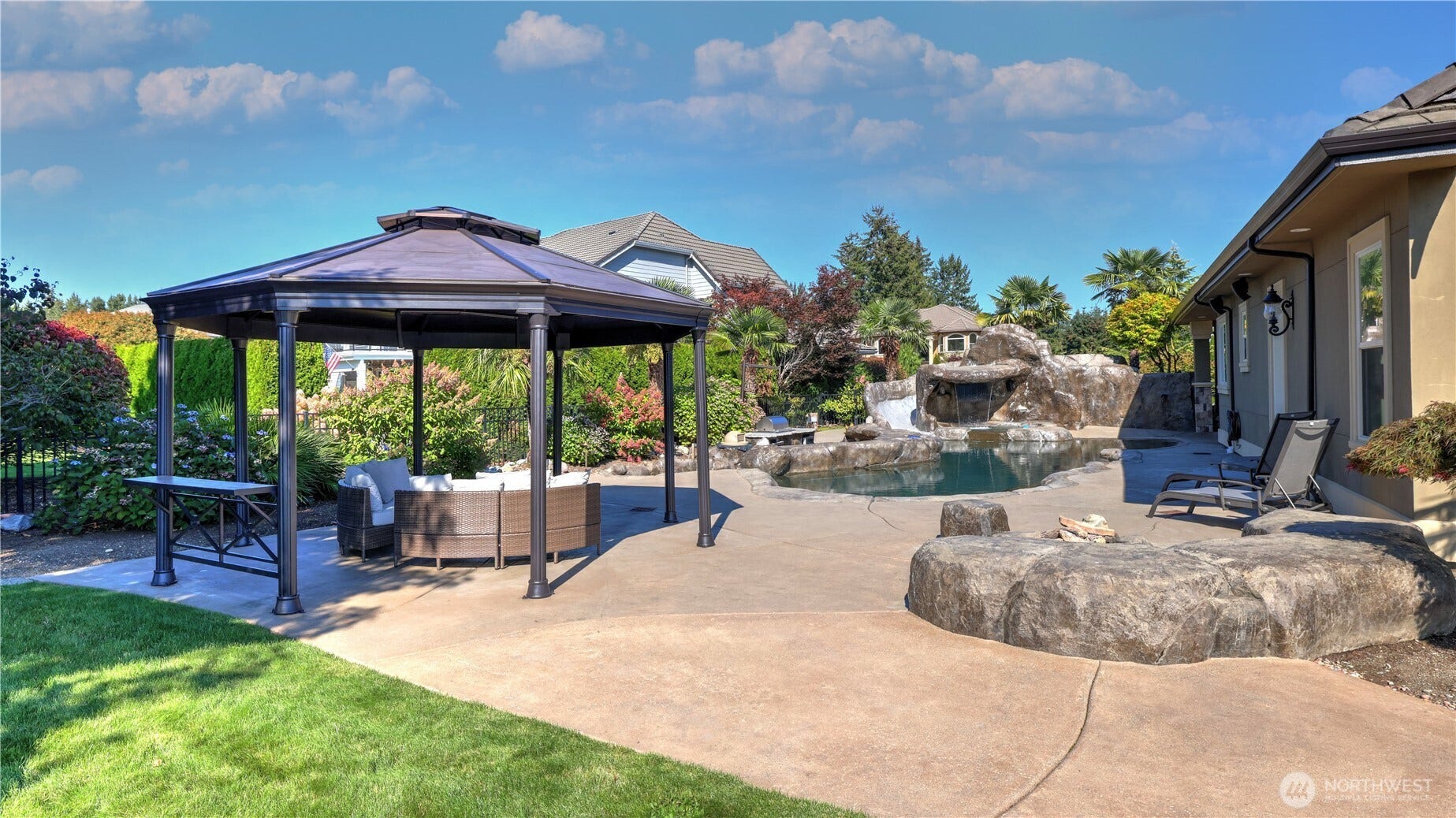 Backyard Oasis with Pool & Gazebo    This beautifully landscaped backyard offers a true resort-style escape, featuring a custom rock pool with waterfall and slide, surrounded by lush greenery and manicured lawn. The spacious patio includes a covered gazebo with comfortable seating, perfect for outdoor dining or relaxing in the shade. Thoughtfully designed for entertaining and leisure, this outdoor retreat blends natural beauty with luxury living.