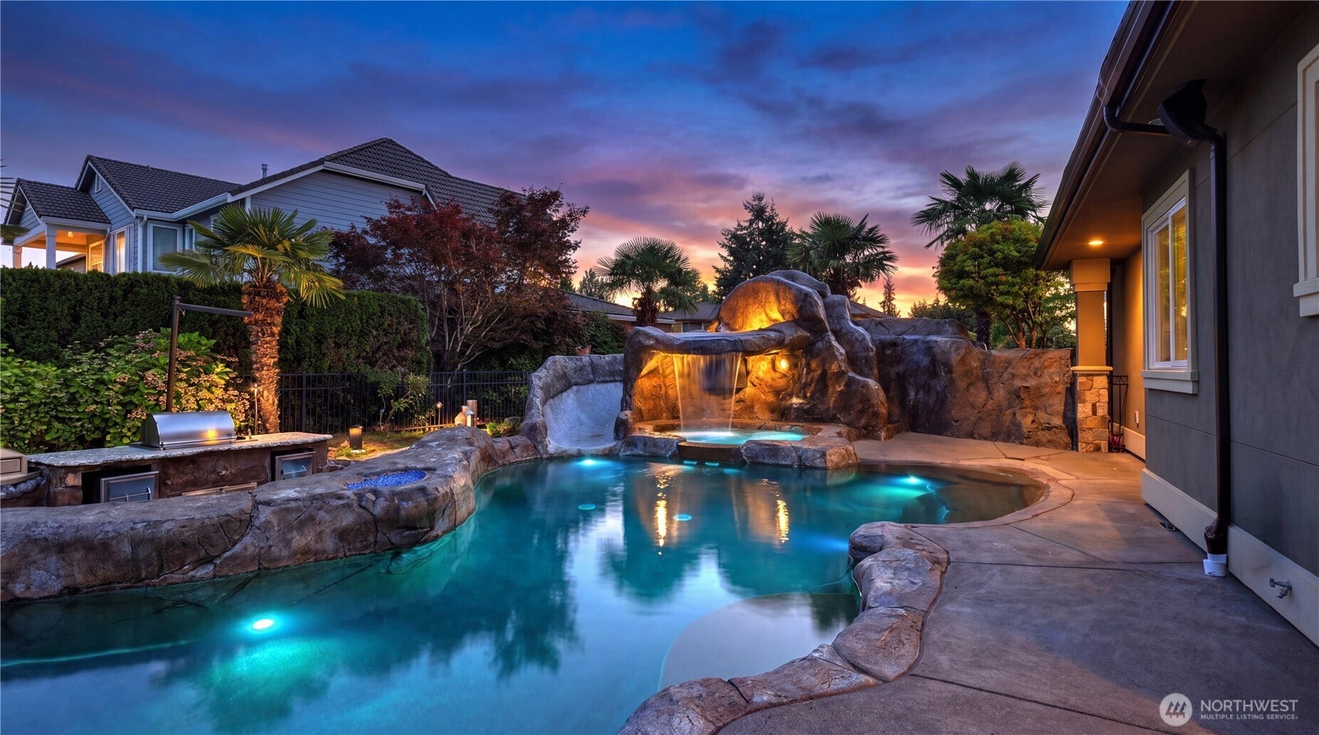 Resort-Style Pool & Outdoor Oasis    This stunning backyard retreat features a custom free-form pool with integrated lighting, a dramatic rock waterfall, and a built-in water slide  all surrounded by natural stonework and lush tropical landscaping. The outdoor kitchen with stainless-steel grill and prep area makes entertaining effortless, while palm trees and sunset lighting create a serene, resort-like ambiance perfect for relaxing or hosting in style.