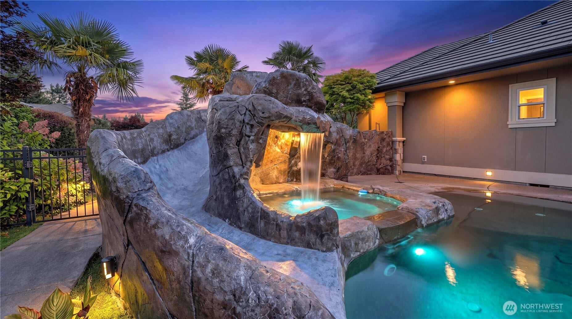 Custom Rock Slide Feature    This resort-style pool is highlighted by a stunning natural rock slide, seamlessly integrated into the grotto design. Crafted for both fun and visual impact, the slide curves gracefully into the illuminated pool below, framed by lush tropical landscaping and a cascading waterfall. Its the perfect centerpiece for an outdoor space that blends luxury, playfulness, and resort-level ambiance.