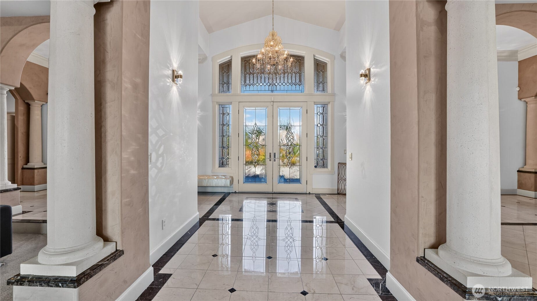 This stunning foyer welcomes guests with architectural drama and timeless elegance. Framed by stately columns and soaring ceilings, the space features polished marble floors with intricate inlays that reflect the light from a dazzling crystal chandelier above. The double glass doors, accented by leaded transom and sidelights, create a luminous entry that floods the area with natural light and offers a glimpse of the manicured grounds beyond. Soft wall sconces add warmth and sparkle, while the symmetrical design and refined finishes set the tone for the homes sophisticated aesthetic. A true statement entry that perfectly balances grandeur and grace.