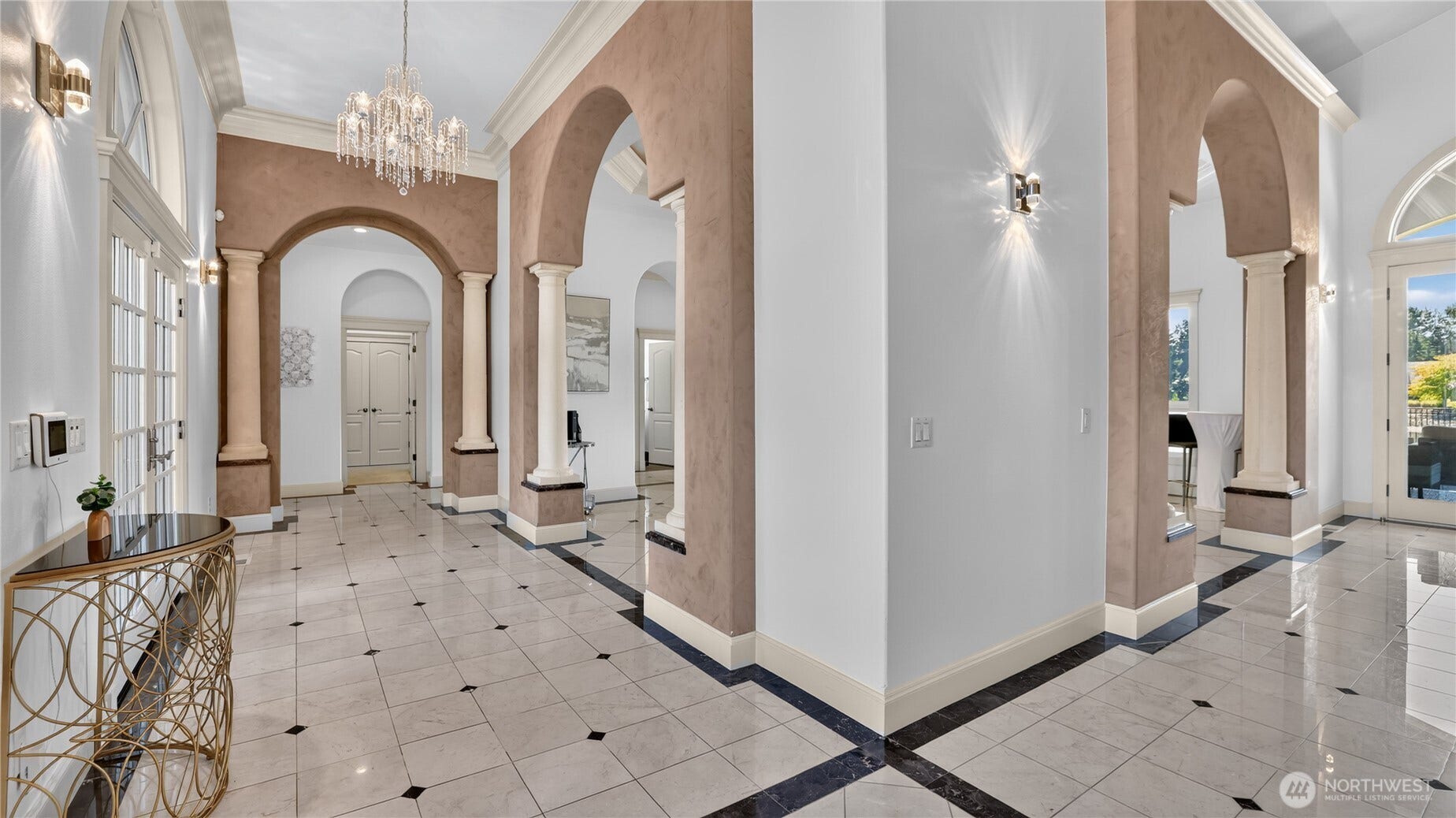 A statement of timeless elegance greets you the moment you step inside. The grand foyer showcases soaring ceilings, graceful archways, and stately columns that frame the space with architectural symmetry. Polished marble floors with contrasting inlays lead the eye toward the formal living areas, while a sparkling crystal chandelier adds a touch of glamour overhead. Soft wall sconces cast a warm glow across the refined palette of cream and taupe, highlighting the craftsmanship and scale of this impressive entry. Designed for both impact and flow, this foyer sets the tone for the homes sophisticated style and resort-like ambiance.