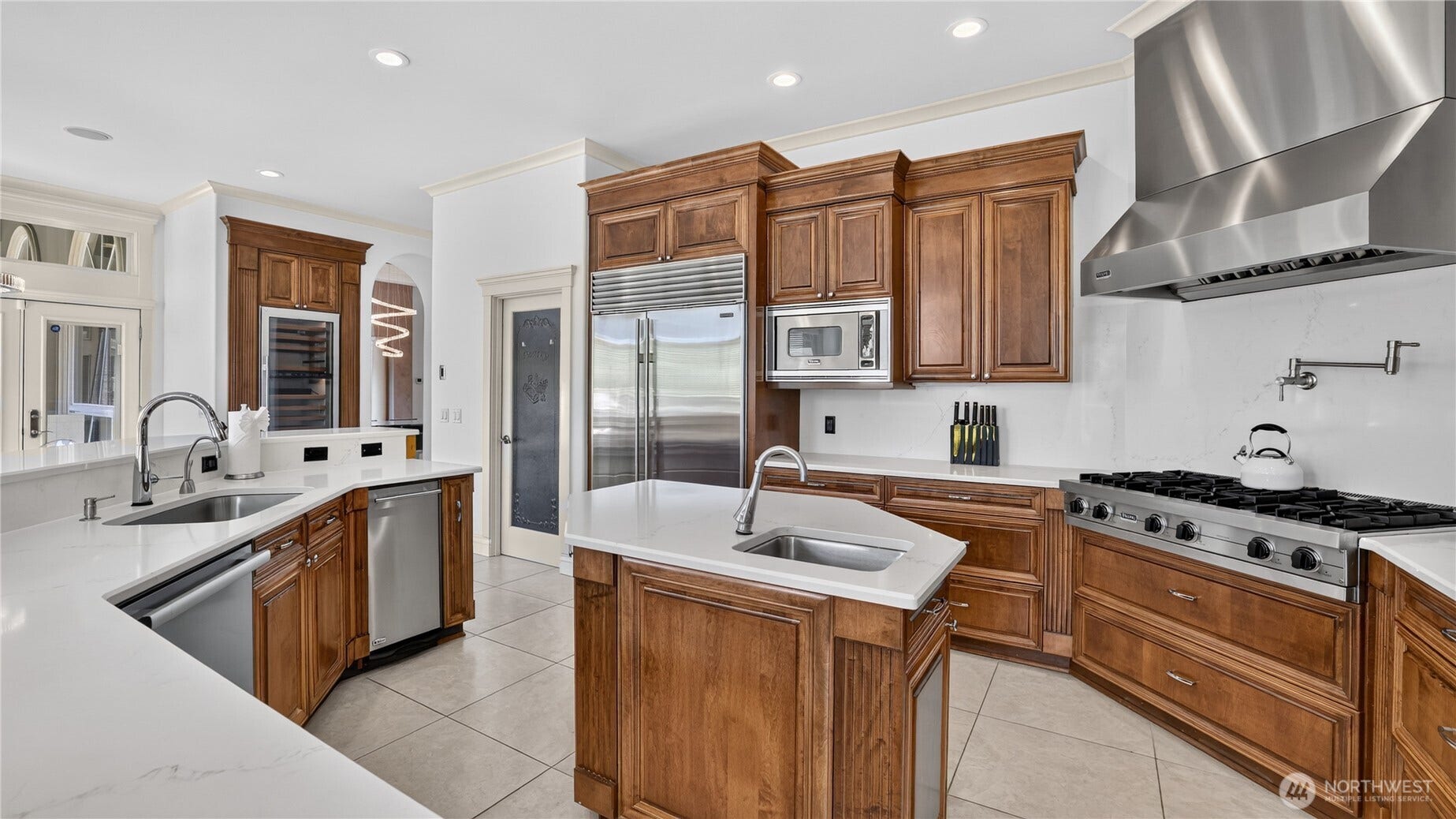 This impressive kitchen is designed for both culinary artistry and everyday luxury. Outfitted with professional-grade Viking appliances, it features a double oven, six-burner gas range, and built-in microwave, all complemented by a Sub-Zero refrigerator for optimal food preservation. A 120-bottle temperature-controlled wine refrigerator anchors the space, perfect for entertaining or fine dining at home. The layout includes a central prep island with sink, pot filler, and expansive workspace counters finished in sleek quartz. Rich wood cabinetry, stainless accents, and recessed lighting complete the sophisticated aesthetic, while the open flow to the adjacent living area ensures seamless connection between cooking and gathering spaces.