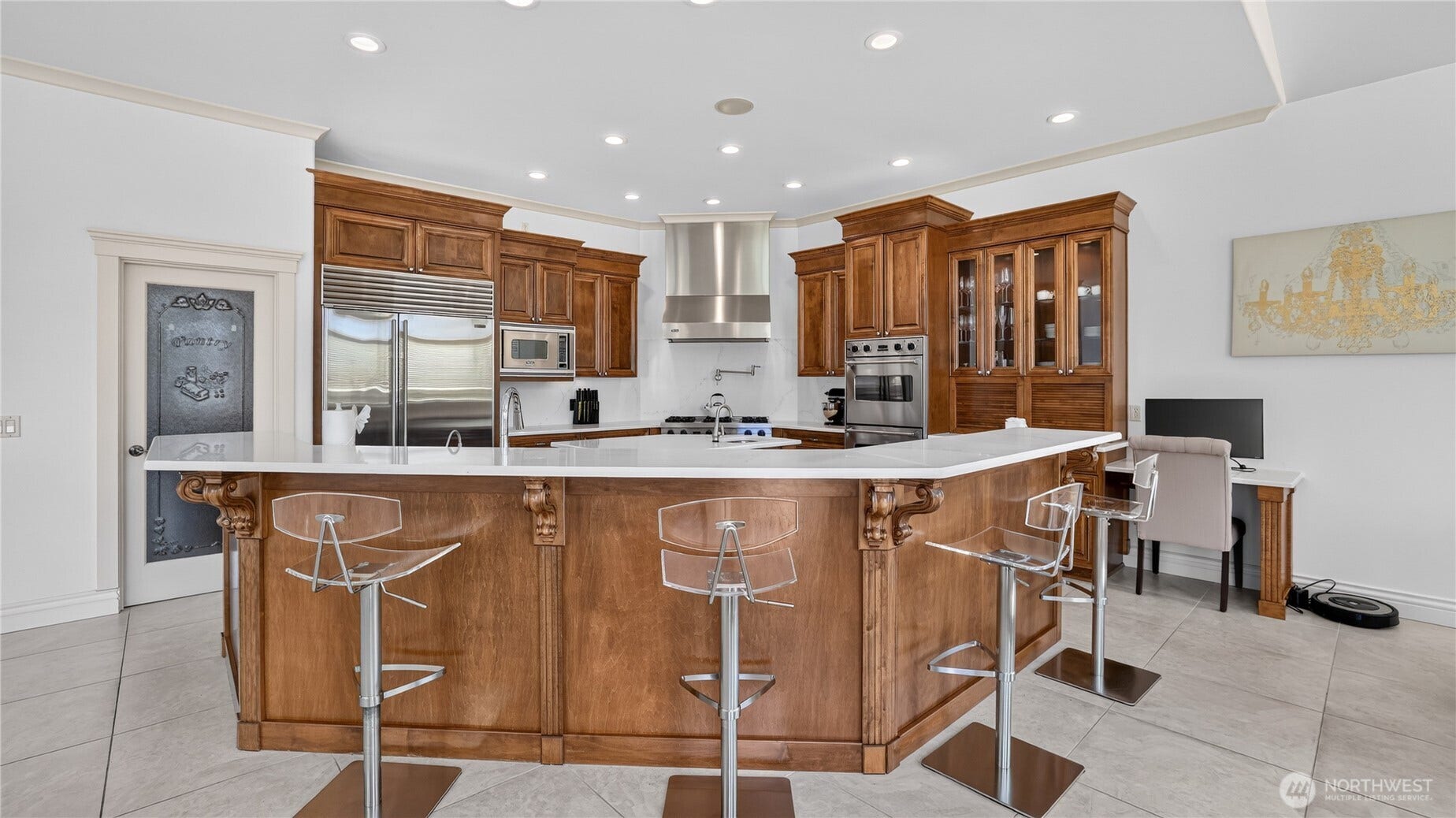 Kitchen    This stunning chefs kitchen combines classic craftsmanship with modern functionality. Rich wood cabinetry and a large center island with quartz countertops create a warm, inviting atmosphere perfect for entertaining. High-end stainless-steel appliances, including double ovens and a built-in refrigerator, complement the elegant design. The space also features a dedicated workstation, ideal for managing daily tasks, all illuminated by recessed lighting that enhances the kitchens refined appeal.