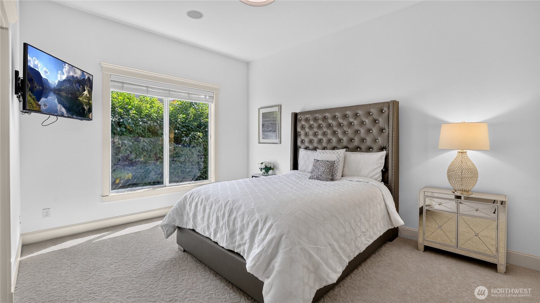 This inviting bedroom offers a peaceful retreat with clean lines and a modern aesthetic. A large window fills the space with natural light and frames lush greenery outside, creating a serene backdrop. The neutral palette, soft carpet, and high ceilings enhance the sense of openness, while the elegant trim and recessed lighting add a refined touch. Designed for comfort and versatility, this room provides an ideal setting for guests or family, maintaining the homes signature sophistication and attention to detail.