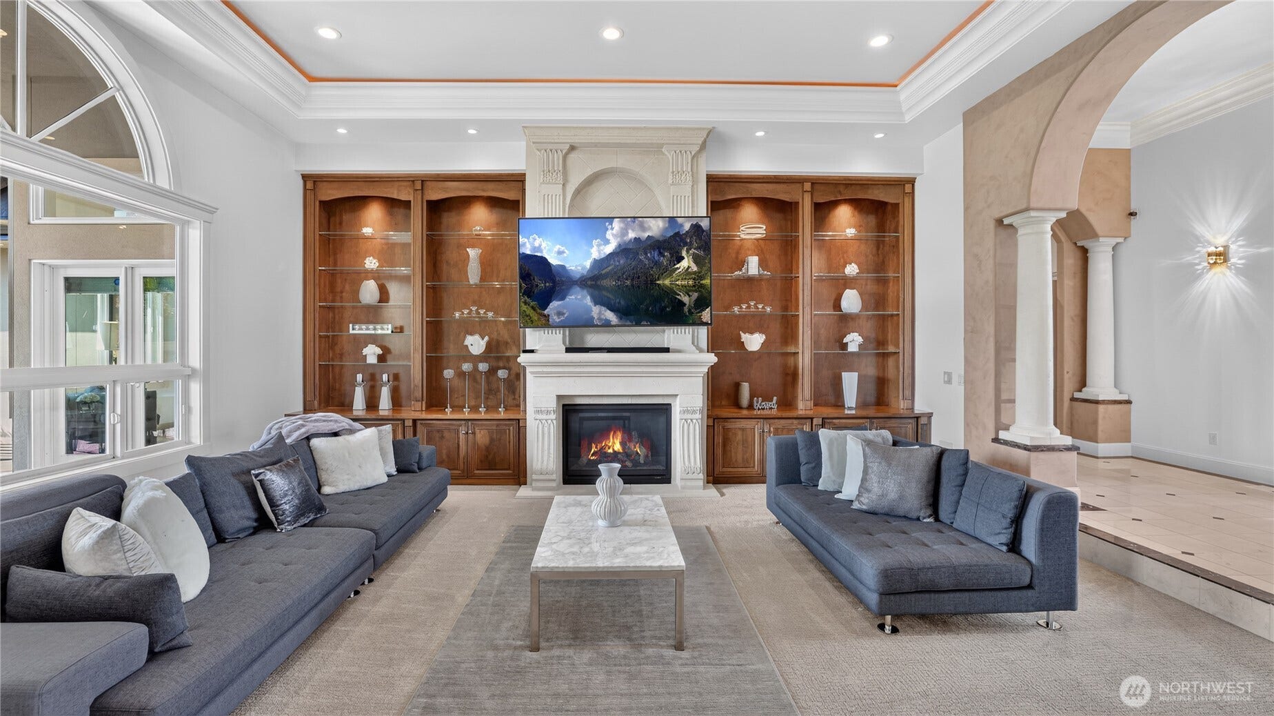 This alternate view of the formal living room highlights its grand symmetry and refined craftsmanship. Centered around an elegant carved stone fireplace, the space is flanked by custom wood built-ins with glass shelving that showcase curated dcor and soft accent lighting. The dual modern sofas create a balanced conversation area anchored by a marble coffee table, while neutral tones and plush textures add warmth and sophistication. Expansive windows and arched openings connect seamlessly to the outdoor living spaces, filling the room with natural light and golf course views. Designed for both comfort and style, this living room embodies timeless luxury and effortless Northwest elegance.