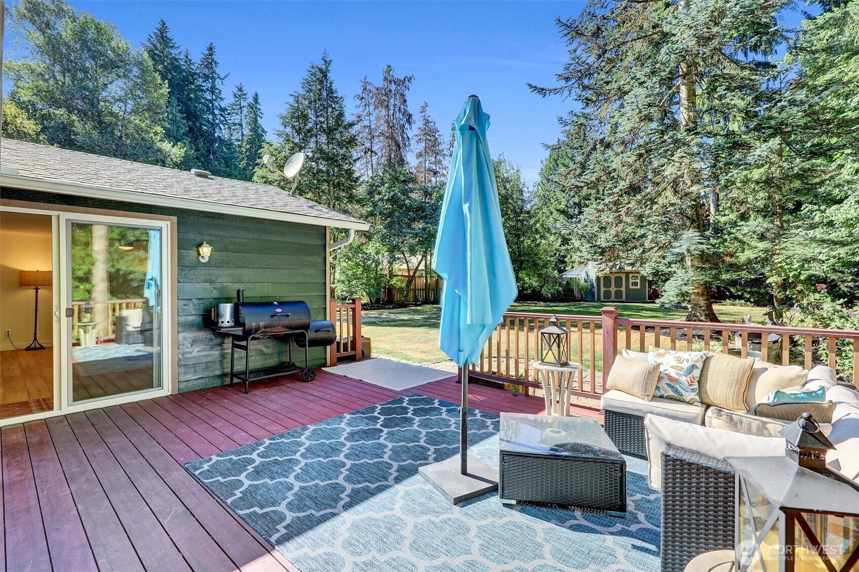 20915 W Richmond Road, Bothell, WA 98021 Spacious backyard deck with outdoor seating, patio umbrella, area rug, sliding glass door, and peaceful wooded surroundings.