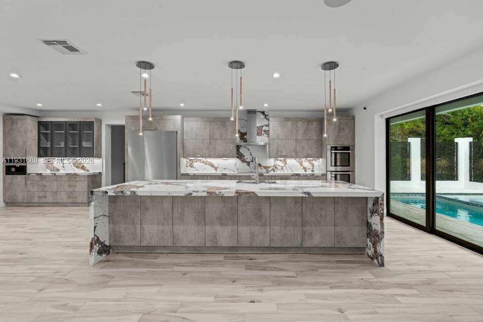 Kitchen featuring custom grey cabinetry, a waterfall-edge stone island with an integrated sink, stone slab backsplash, stainless steel built-in appliances, and wood-finish flooring