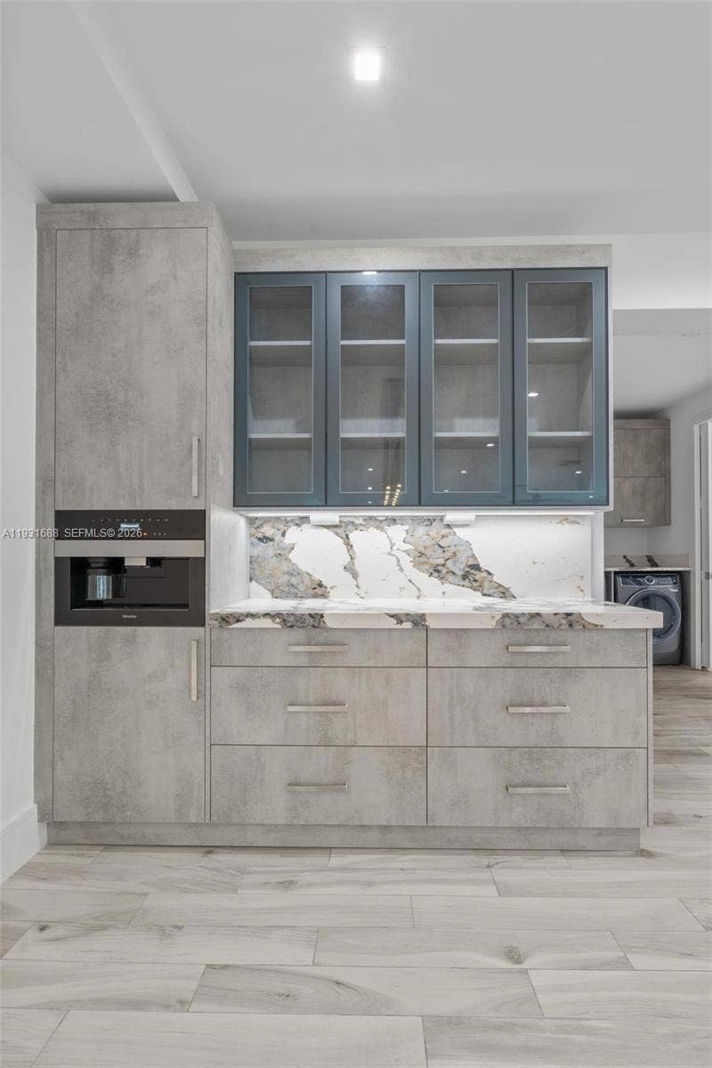 Custom cabinetry featuring grey textured finish and glass-front upper storage, stone-patterned backsplash, quartz countertop, and integrated built-in coffee station