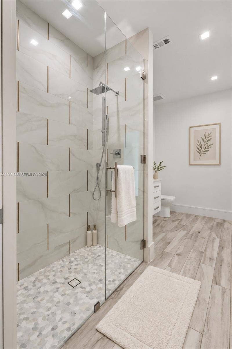 Walk-in shower featuring large-format tile walls with metallic inlay, river stone tile flooring, frameless glass enclosure, rainfall shower head, and wood-finish flooring
