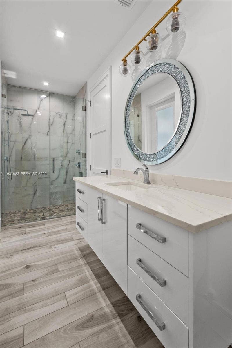 White high-gloss cabinetry with a stone countertop, walk-in shower with floor-to-ceiling tile, circular mirror with decorative frame, brushed-gold finish vanity lighting, and wood-finish flooring