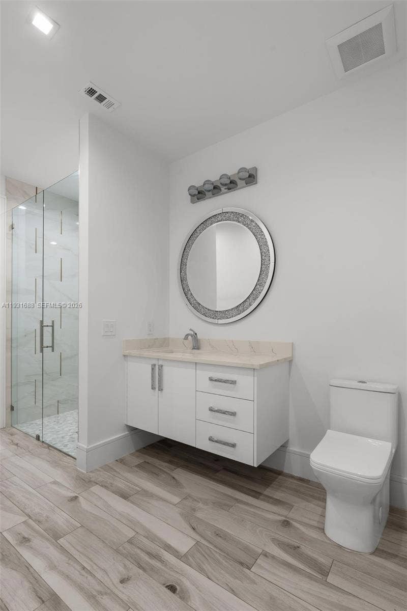 Floating white vanity with stone-finish countertop, wood-finish flooring, glass-enclosed shower with tiled walls, and polished hardware