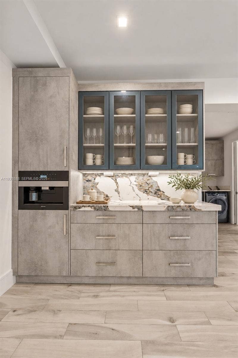 Textured grey cabinetry featuring glass-front upper displays, stone-patterned backsplash and countertop, integrated coffee system, and wood-finish tile flooring