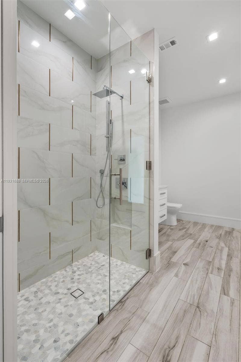 Walk-in shower featuring large-format wall tile with metallic inlay, river stone mosaic floor, frameless glass enclosure, and rainfall showerhead, complemented by wood-finish flooring