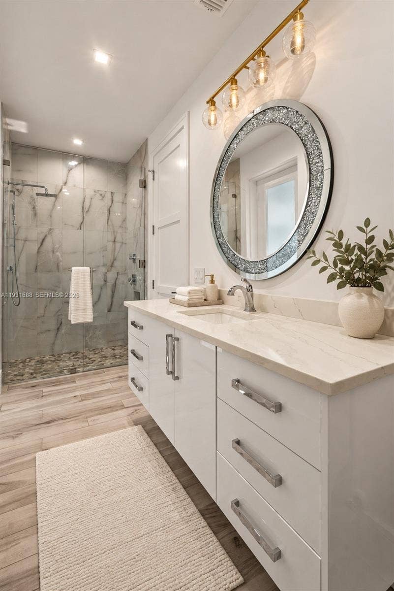 Bathroom featuring a white gloss vanity with stone-finish countertop, walk-in shower with stone tile surround and pebble floor, wood-finish flooring, and wall-mounted multi-light fixture