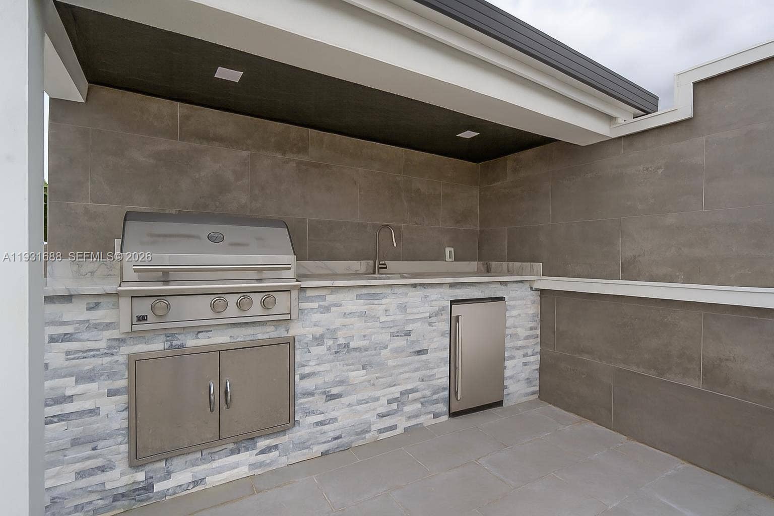 Outdoor kitchen featuring stainless steel grill, under-counter refrigeration, stone-veneer cabinetry base, and tile backsplash
