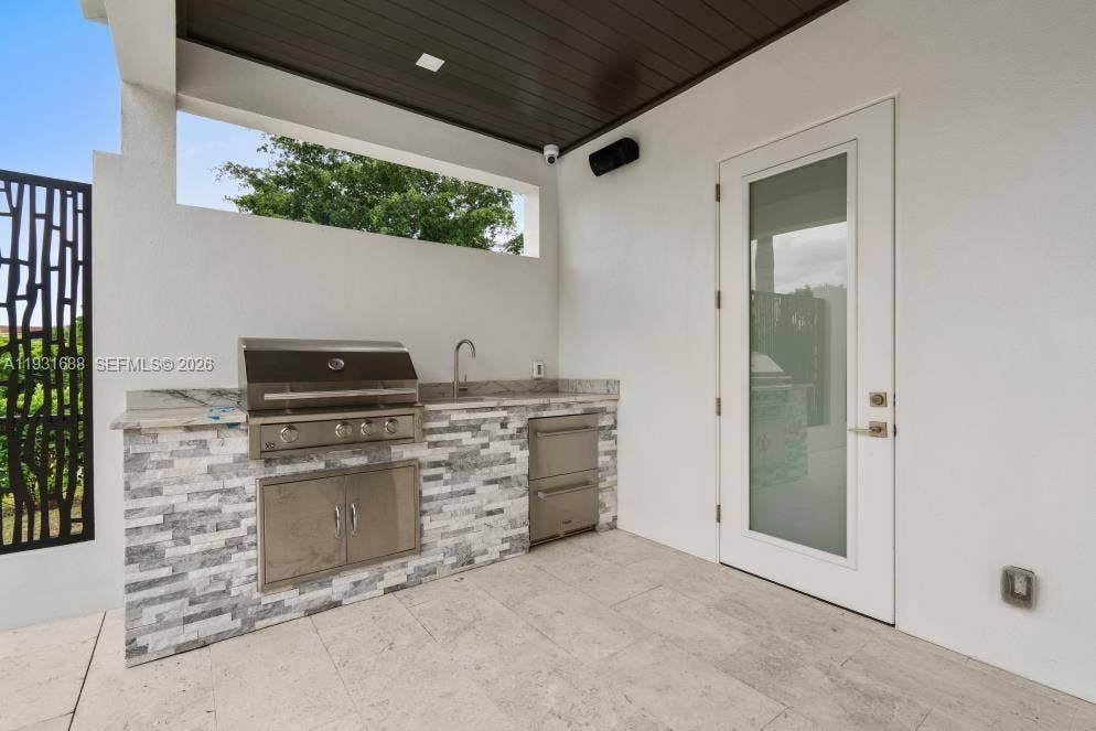 Covered outdoor kitchen featuring stone-veneer cabinetry, stone countertops, a stainless steel built-in grill, an under-counter refrigerator, and a stainless steel sink