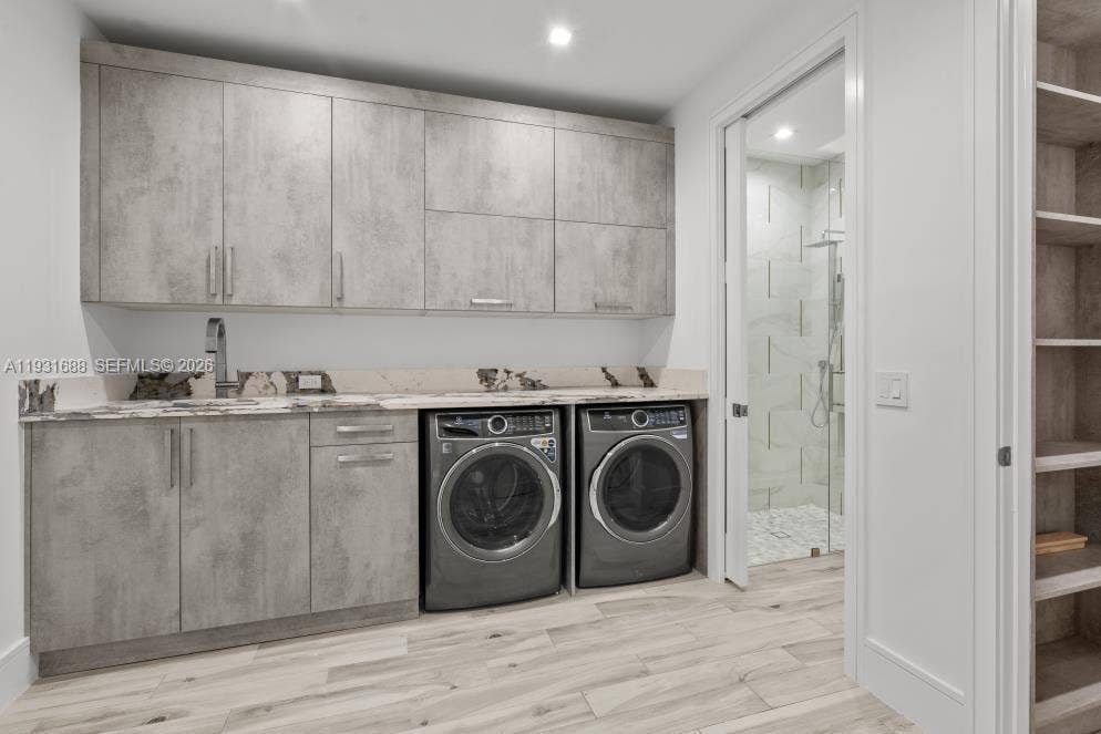 Laundry room featuring grey-toned cabinetry, stone-surface countertop with integrated sink, wood-finish flooring, and built-in shelving, with a glass-enclosed shower stall visible through the doorway