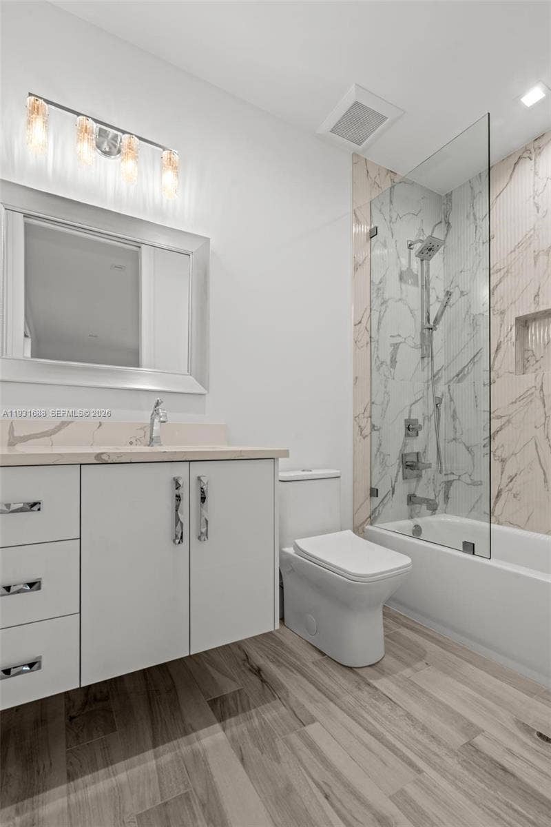 Bathroom featuring a wood-finish floor, white vanity with stone-surface countertop, full-height decorative tile shower surround with a glass panel, and polished chrome fixtures