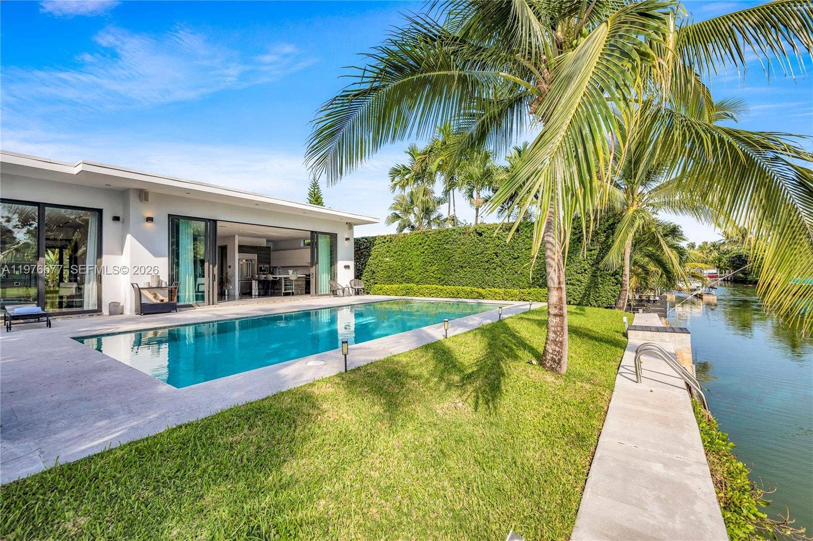 Waterfront property featuring a rectangular in-ground pool, concrete patio, manicured lawn, and mature palm trees