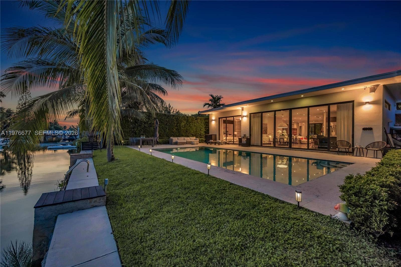 Waterfront exterior featuring a rectangular swimming pool, concrete patio, glass wall panels, and a concrete seawall with wood-finish decking