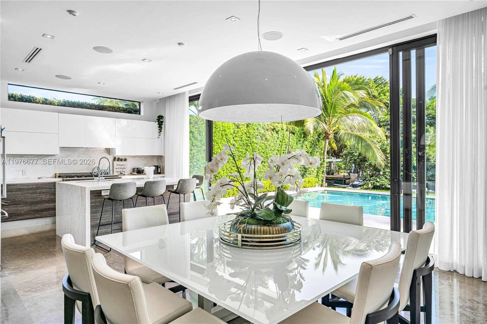 Open-concept kitchen and dining area featuring a central island with stone-finish waterfall edge, high-gloss flat-panel cabinetry, recessed lighting, and expansive floor-to-ceiling glass sliding doors opening to a swimming pool
