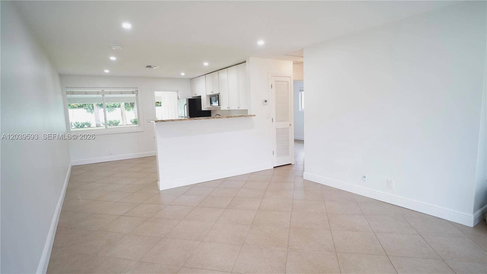 Open-concept interior featuring light-colored tile flooring, recessed ceiling lighting, a kitchen peninsula with stone-finish countertops, and white cabinetry
