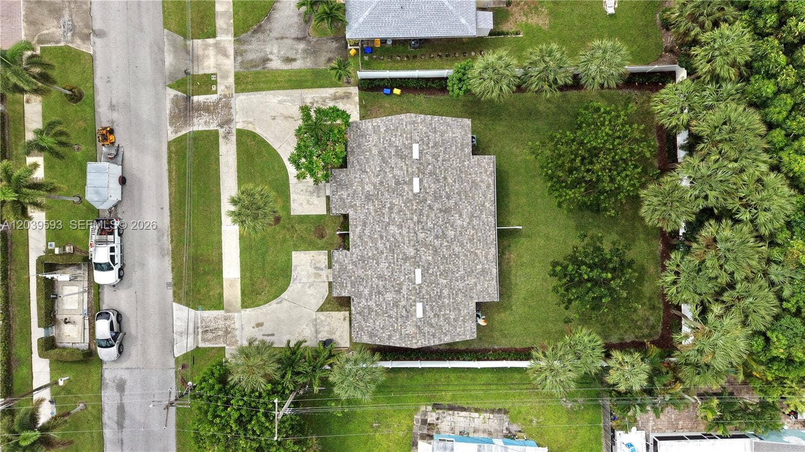 Aerial view featuring a paved circular driveway, a shingle-style roof, and a fenced-in lawn with mature landscaping