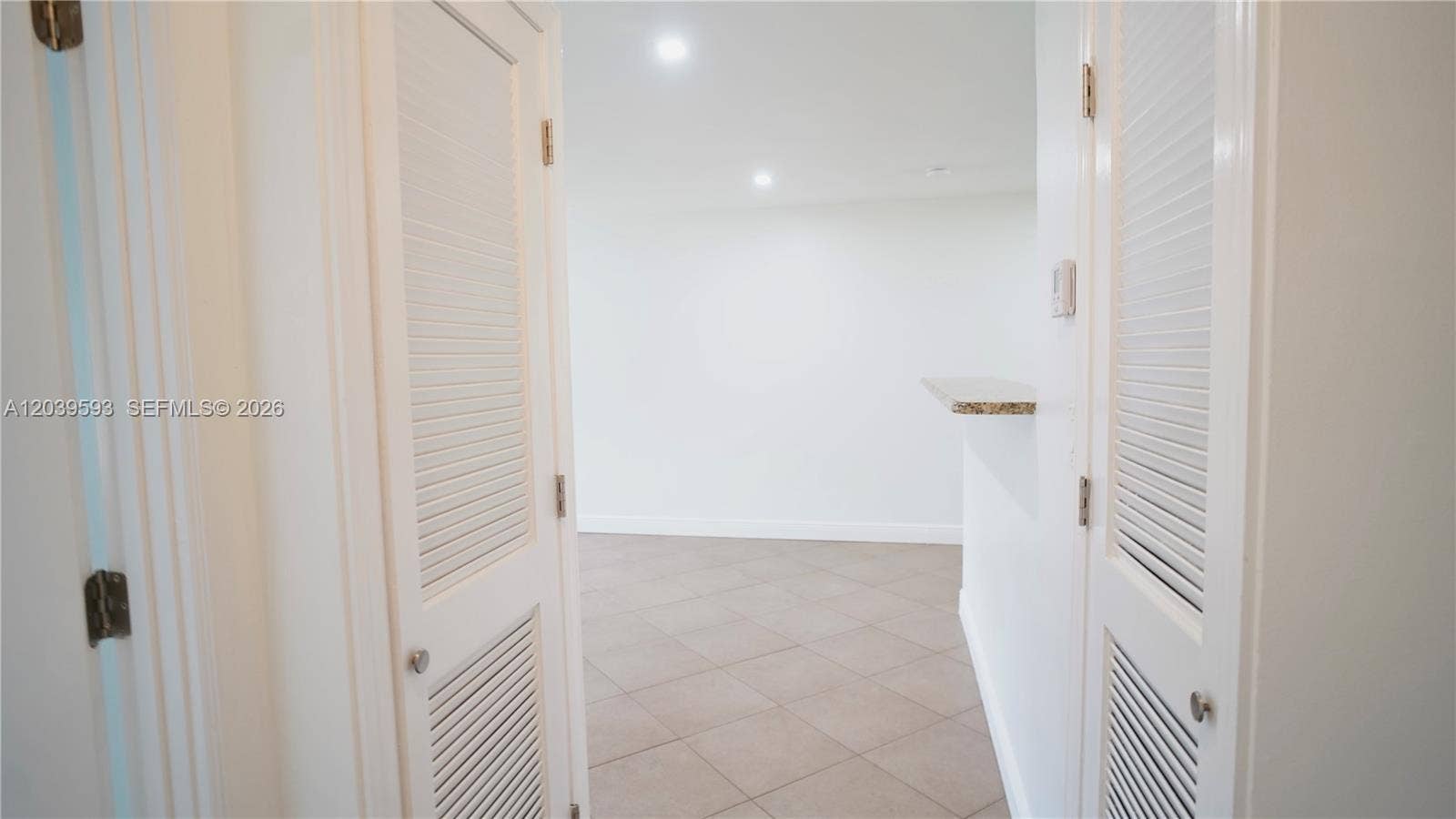 Dual louvered closet doors, neutral tile flooring, recessed ceiling lighting, and a stone-surface pass-through ledge