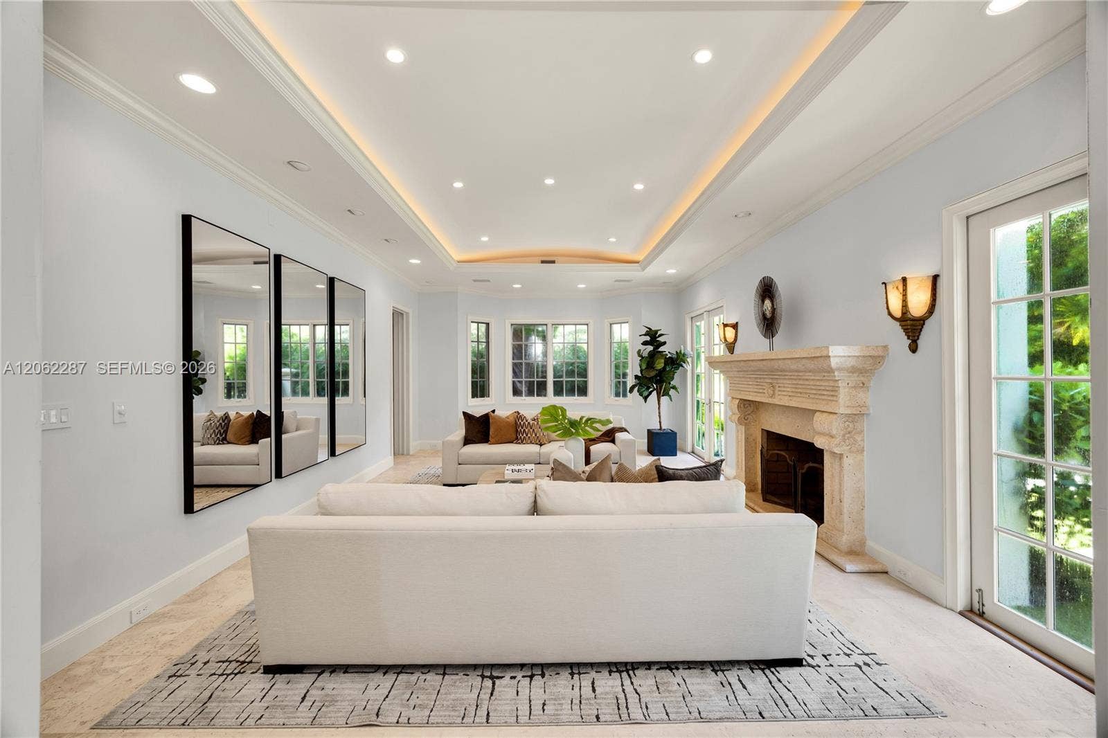 Living area featuring a tray ceiling with recessed lighting, carved stone fireplace mantel, wall-to-wall mirrors, light-toned stone flooring, and multiple sets of glass-paned doors