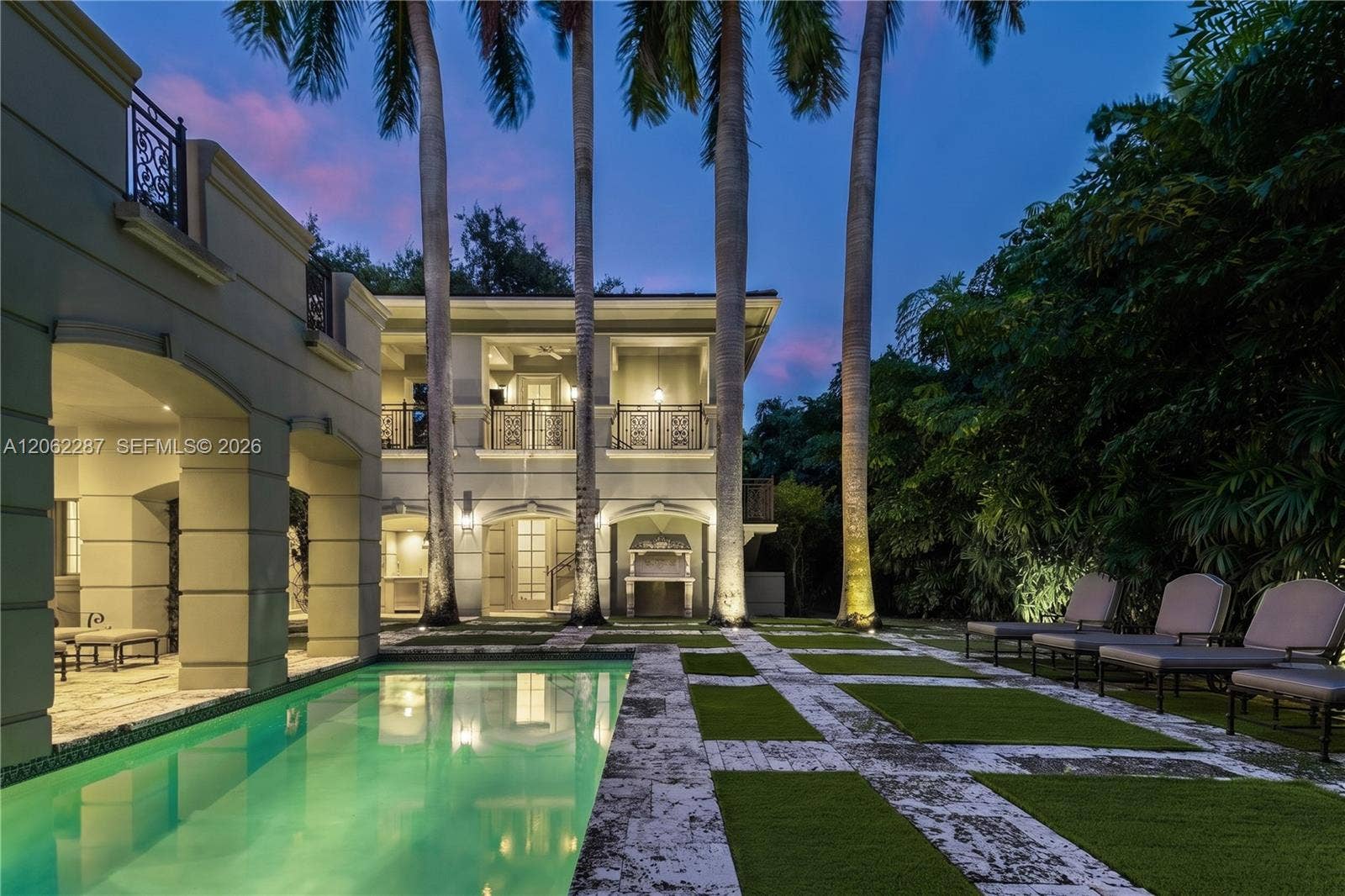 Exterior space featuring a rectangular swimming pool, stone-paver hardscaping, grid-patterned turf landscaping, mature palm trees, and two-story structures with arched loggias and wrought-iron balcony railings