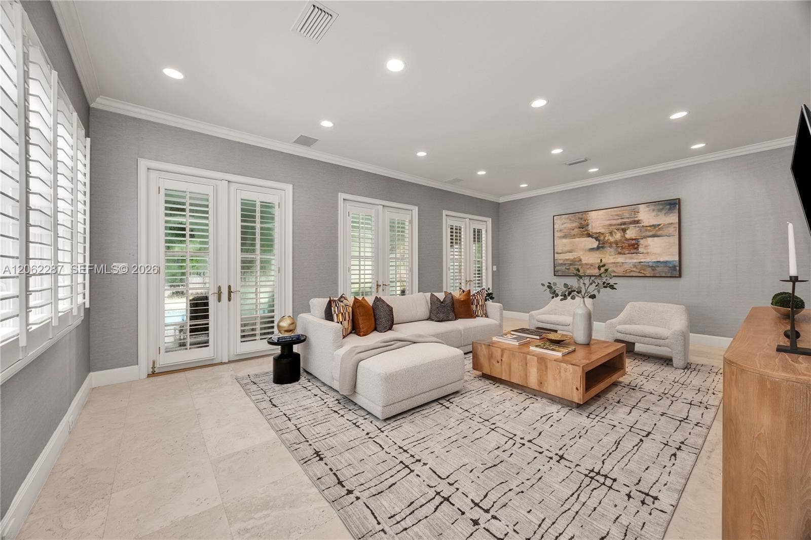 Neutral-toned interior space featuring light-colored stone flooring, recessed ceiling lighting, crown molding, and dual glass-paned French doors with interior shutters
